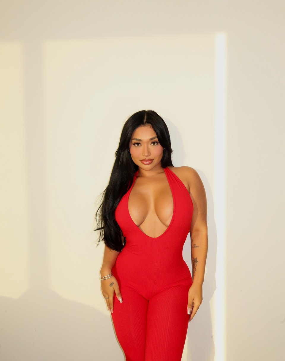 Ale V in a vivid red jumpsuit against a pale wall.